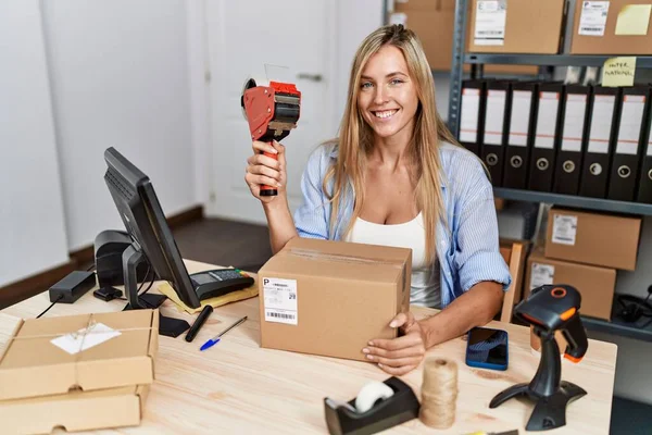 Young blonde woman ecommerce business worker holding packing tape machine at office