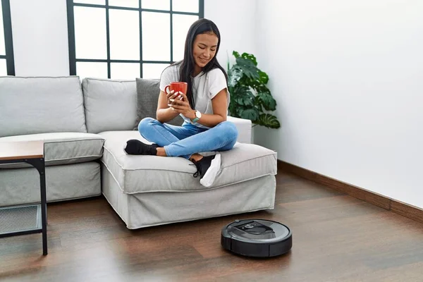 Genç Latin kadın evde akıllı robot süpürgesi kullanarak kahve içti.