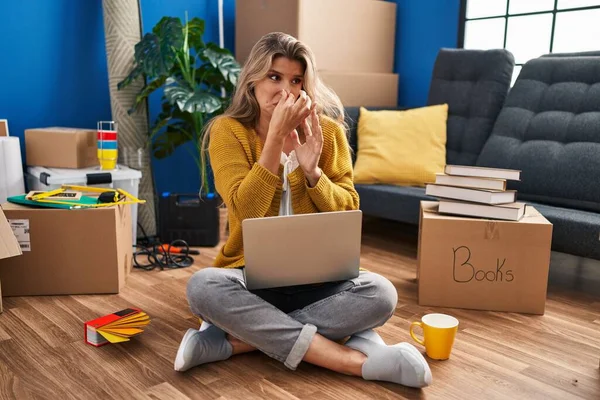 Young woman sitting on the floor at new home using laptop smelling something stinky and disgusting, intolerable smell, holding breath with fingers on nose. bad smell