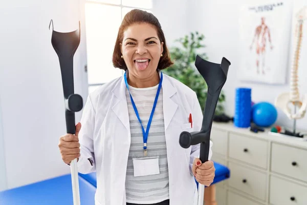 Middle Age Hispanic Physiotherapy Woman Holding Crutches Sticking Tongue Out —  Fotos de Stock