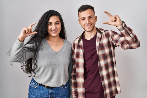 Young hispanic couple standing over white background smiling and confident gesturing with hand doing small size sign with fingers looking and the camera. measure concept.