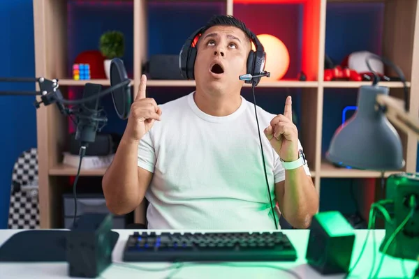 Молодой Латиноамериканец Играющий Видеоигры Поражен Удивлен Глядя Вверх Показывая Пальцами — стоковое фото