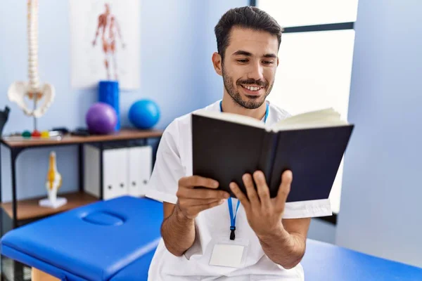Young Hispanic Man Wearing Physiotherapist Uniform Reading Book Rehab Clinic — ストック写真