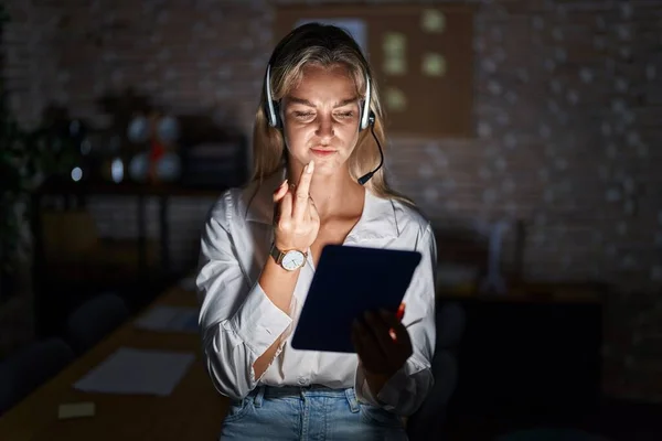 Young blonde woman working at the office at night showing middle finger, impolite and rude fuck off expression