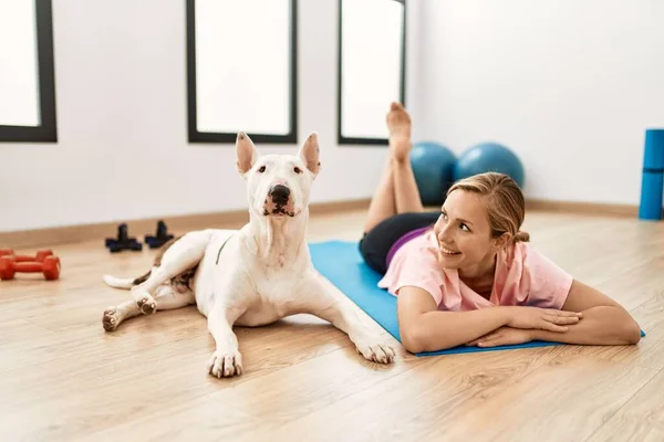 Young caucasian woman smiling confident lying on yoga mat with dog at sport center