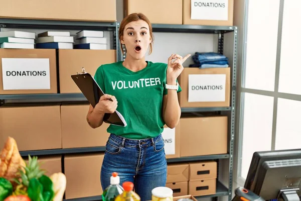 Young Brunette Woman Wearing Volunteer Shirt Donations Stand Surprised Pointing — Stock Photo, Image