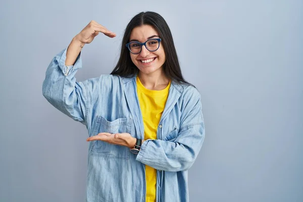 Young hispanic woman standing over blue background gesturing with hands showing big and large size sign, measure symbol. smiling looking at the camera. measuring concept.