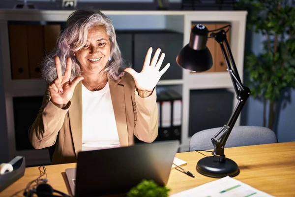 白发苍苍的中年妇女深夜使用电脑笔记本电脑工作 带着八号手指 面带微笑 自信而快乐 — 图库照片