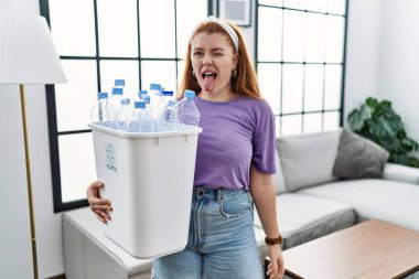Genç kızıl saçlı kadın elinde plastik şişelerle çöp sepetini tutarak komik bir ifadeyle dilini dışarı çıkartıyor. duygu kavramı. 
