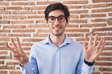 Young hispanic man standing over brick wall background showing and pointing up with fingers number eight while smiling confident and happy. 