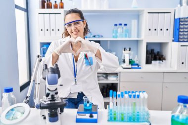 Young hispanic woman working at scientist laboratory smiling in love doing heart symbol shape with hands. romantic concept. 