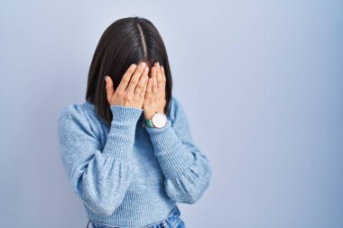 Mavi arka planda duran genç İspanyol kadın. Ağlarken suratını ellerle kapatan üzgün bir ifade. depresyon kavramı. 