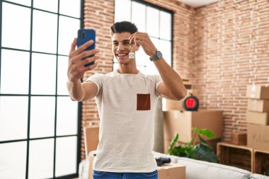Young hispanic man having video call holding key of house at new home
