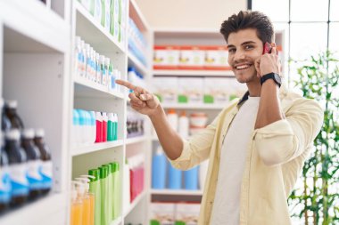 Genç İspanyol adam gülümseyerek eczanede akıllı telefondan konuşuyor.