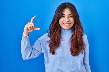 Hispanic young woman standing over blue background smiling and confident gesturing with hand doing small size sign with fingers looking and the camera. measure concept. 