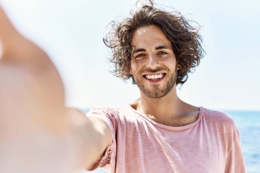 Mutlu gülümseyen genç İspanyol adam sahilde kameranın önünde selfie çekiyor..