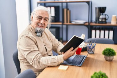 Middle age grey-haired man business worker using laptop reading book at office