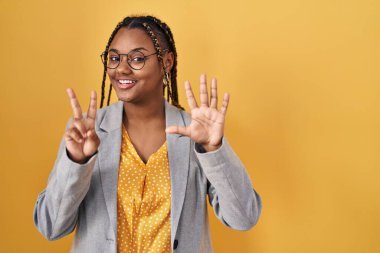 African american woman with braids standing over yellow background showing and pointing up with fingers number seven while smiling confident and happy. 