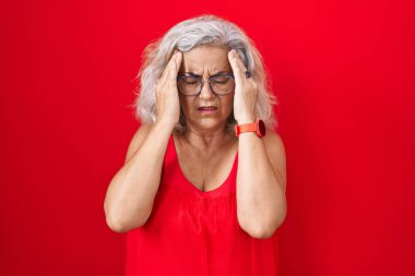 Orta yaşlı, gri saçlı bir kadın. Kırmızı arka planda duruyor. Elleri başının üstünde, stres yüzünden başı ağrıyor. migren ağrısı. 