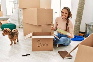 Genç, güzel, İspanyol bir kadın yeni evinde köpekle birlikte karton kutuları açıyor.