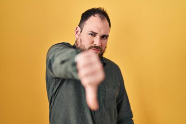 Plus size hispanic man with beard standing over yellow background looking unhappy and angry showing rejection and negative with thumbs down gesture. bad expression. 