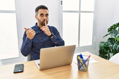 Ofiste sakallı, laptopla çalışan genç bir İspanyol adam elleriyle dudaklarını gösteren parmaklarıyla sessiz olmak istiyor. sessizlik ve gizli konsept. 
