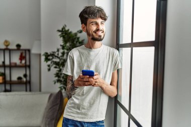 Genç İspanyol adam evde akıllı telefon kullanarak kendinden emin gülümsüyor.