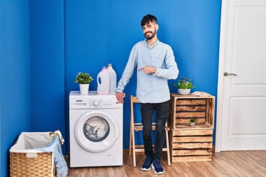 Çamaşır odasında çamaşır yıkayan, sakallı, genç, İspanyol bir adam elini ve parmağını göstererek mutlu bir şekilde duruyor. 