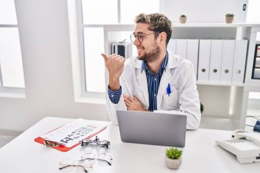 Hispanic man with beard wearing doctor uniform and stethoscope pointing thumb up to the side smiling happy with open mouth 
