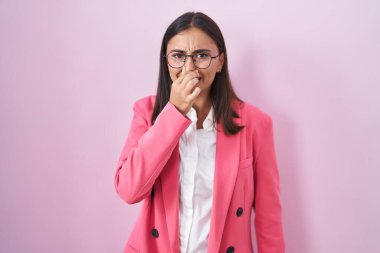 Young hispanic woman wearing business clothes and glasses smelling something stinky and disgusting, intolerable smell, holding breath with fingers on nose. bad smell 