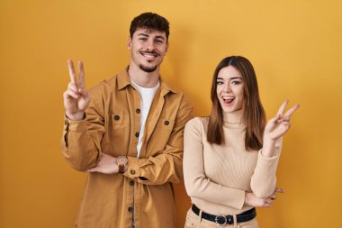 Young hispanic couple standing over yellow background smiling with happy face winking at the camera doing victory sign with fingers. number two. 