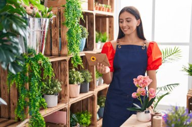 Young beautiful hispanic woman florist holding envelope letter and plant at florist