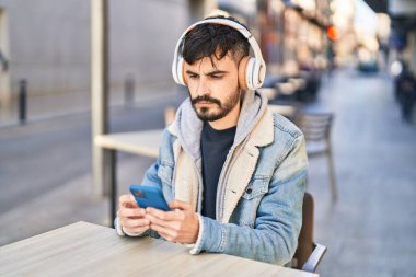 Young hispanic man listening to music sitting on table at coffee shop terrace