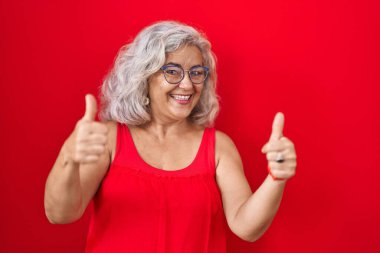 Kırmızı arka planda duran gri saçlı orta yaşlı kadın elinde olumlu bir jest yapıyor, gülümsüyor ve mutlu. Neşeli ifade ve kazanan jest. 