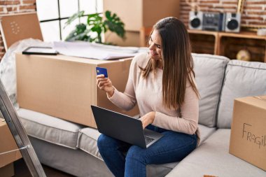 Young beautiful hispanic woman using laptop and credit card sitting on sofa at new home