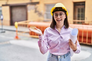 Hispanic girl wearing architect hardhat at construction site celebrating achievement with happy smile and winner expression with raised hand 