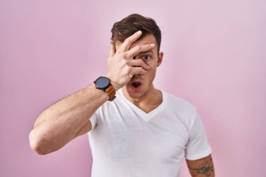 Caucasian man standing over pink background peeking in shock covering face and eyes with hand, looking through fingers with embarrassed expression. 