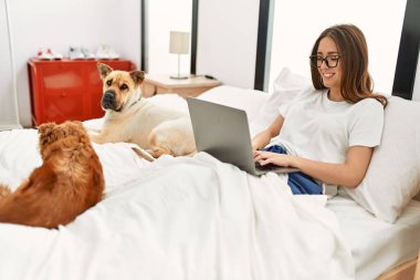 Genç İspanyol kadın, yatak odasında köpeklerle birlikte dizüstü bilgisayar kullanıyor.