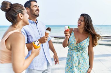 Üç genç İspanyol arkadaş gülümseyerek kumsalda dondurma yiyor..