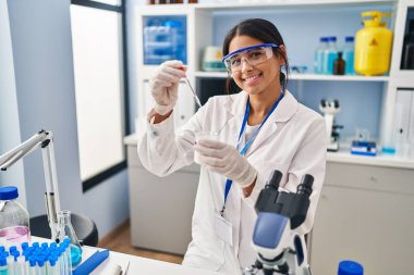 Laboratuvarda pipet kullanan genç Latin kadın bilim adamı üniforması giyiyor.