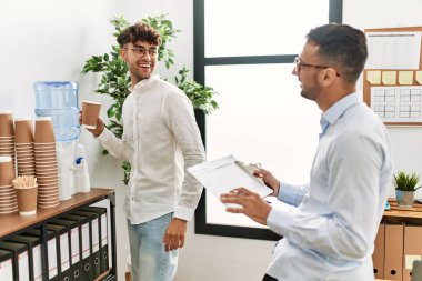 Two hispanic men business workers drinking water and writing on clipboard working at office