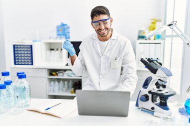 Laboratuvarda çalışan genç İspanyol bir adam dizüstü bilgisayarla gururla bağırıyor, zaferi kutluyor ve başarıyı kollarını kaldırarak kutluyor. 