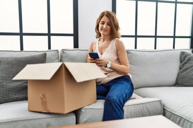 Orta yaşlı bir kadın koltukta oturmuş telefonda teslimat paketini kontrol ediyor. Pozitif ve mutlu görünüyor. Kendine güvenen bir gülümsemeyle dişlerini gösteriyor. 