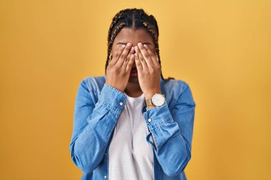 African american woman with braids standing over yellow background rubbing eyes for fatigue and headache, sleepy and tired expression. vision problem 