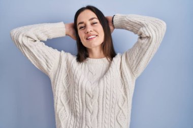 Genç esmer kadın mavi arka planda duruyor rahatlıyor ve esniyor, kolları ve elleri başının arkasında mutlu bir şekilde gülümsüyor. 