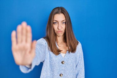 Young woman standing over blue background doing stop sing with palm of the hand. warning expression with negative and serious gesture on the face. 