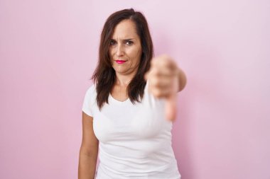 Middle age brunette woman standing over pink background looking unhappy and angry showing rejection and negative with thumbs down gesture. bad expression. 