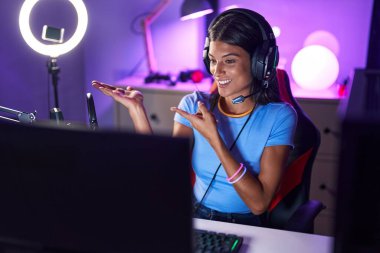 Brunette young woman playing video games amazed and smiling to the camera while presenting with hand and pointing with finger. 