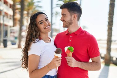 Young hispanic couple tourists hugging each other eating ice cream at seaside