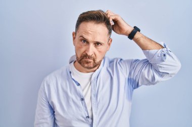 Orta yaşlı, beyaz bir adam mavi arka planda duruyor. Kafası karışmış ve soru soruyor. Şüpheyle kararsız, kafa kafaya düşünerek. Düşünceli kavram. 